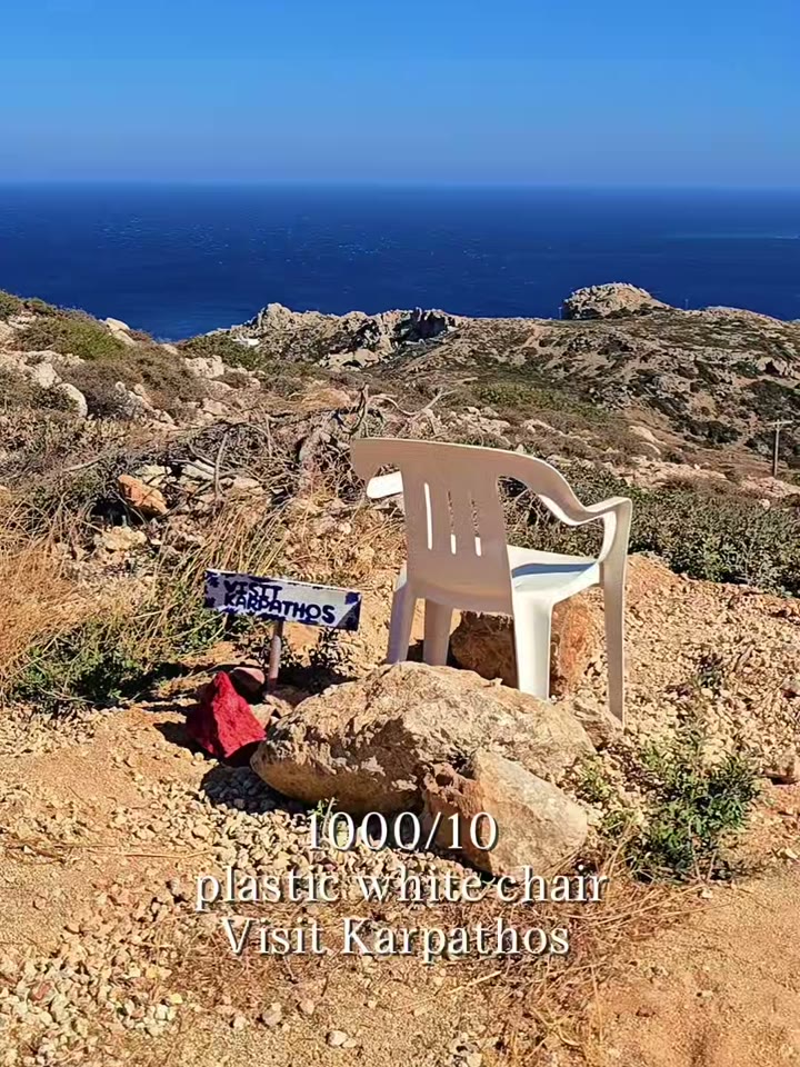 A white plastic chair on a cliff of Karpathos, facing the Aegean, next to a Visit Karpathos sign.